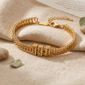 May include: Gold-colored bracelet with a woven chain design. The bracelet features three rectangular charms engraved with the names "DANIEL", "SOPHIA", and "DIANA". The bracelet is displayed on a beige fabric surface.