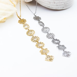May include: Two necklaces with chakra symbols. One is gold-toned, the other silver-toned. Each necklace features a chain with seven pendant charms, each representing a chakra. The pendants are geometric shapes with intricate cut-out designs.