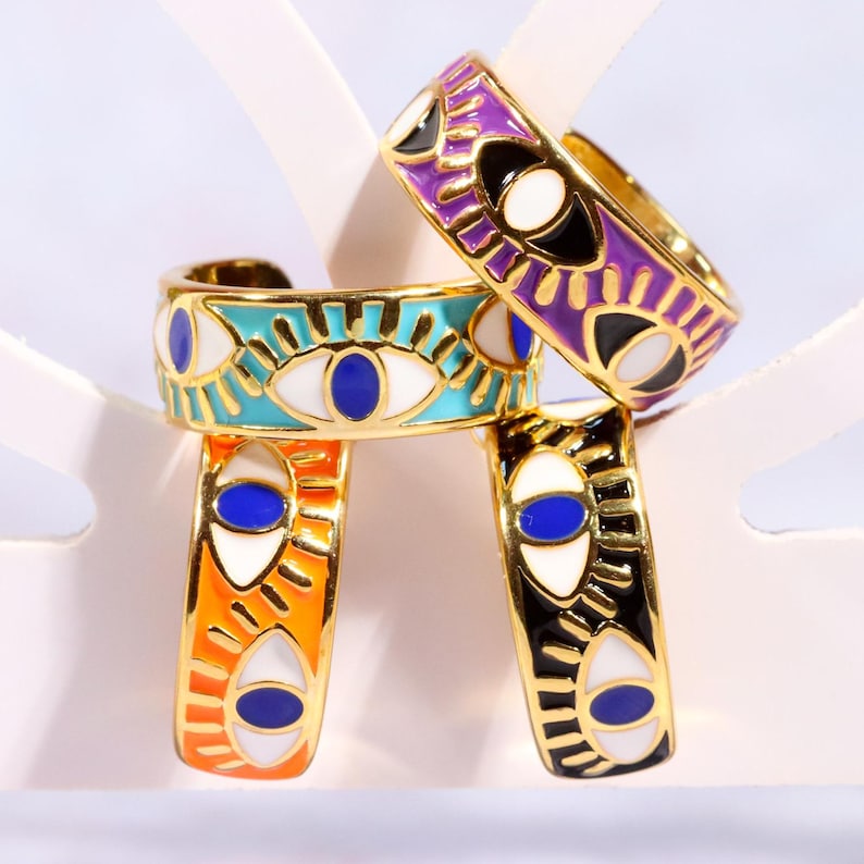 Puede incluir: Cuatro anillos ajustables dorados con dise&ntilde;os de ojos esmaltados. Los anillos presentan varios colores, incluyendo turquesa, naranja, negro y morado, con detalles de ojos blancos y azules. Los anillos est&aacute;n apilados, mostrando sus intrincados dise&ntilde;os.