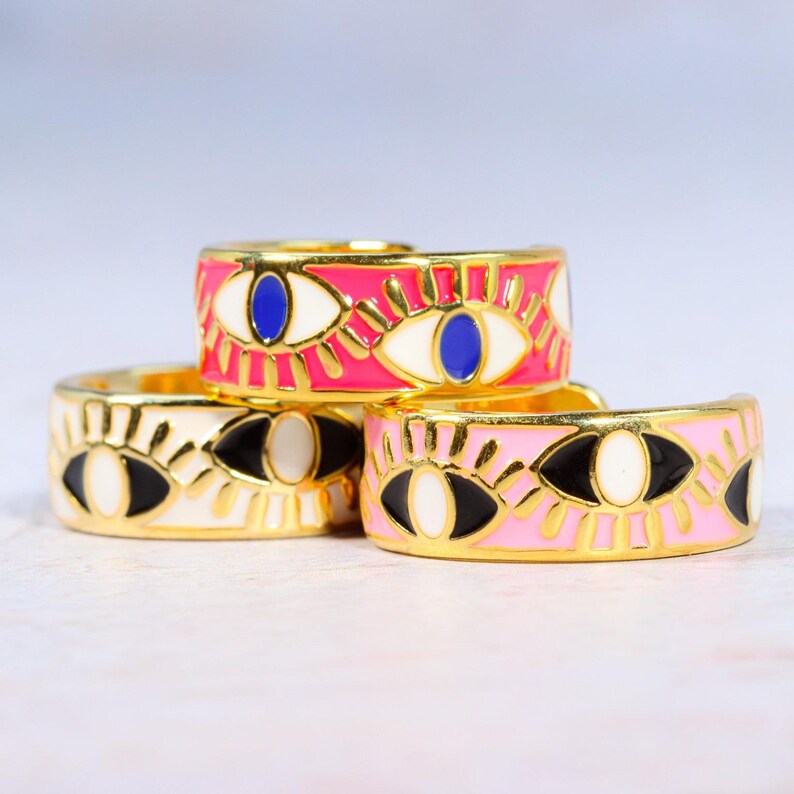 Puede incluir: Tres anillos dorados con un dise&ntilde;o de ojo. Un anillo es rosa con un ojo azul, otro es blanco con un ojo negro y el tercero es rosa con un ojo negro. Los anillos est&aacute;n apilados.