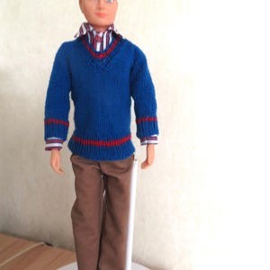 May include: A doll with dark hair and blue eyes wearing a blue V-neck jumper with red stripes on the cuffs and hem, a striped collared shirt underneath, and brown trousers. The doll is standing upright on a white circular base with a support stand.