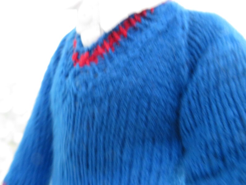 May include: A blue knitted jumper featuring a V-neckline with a red stitched trim detail. The garment is crafted with a ribbed texture throughout the body and sleeves, and it is worn over a white collared shirt visible at the neckline.