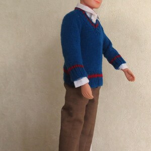 May include: A doll with dark hair and blue eyes wearing a white collared shirt under a blue V-neck sweater with red stripes on the cuffs and hem. The doll is dressed in brown trousers and dark brown shoes, standing on a white circular base.