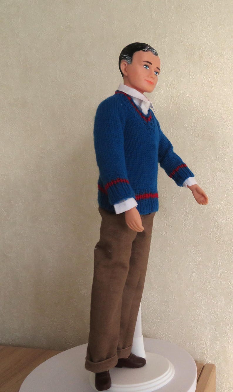 May include: A doll with dark hair and blue eyes wearing a white collared shirt under a blue V-neck sweater with red stripes on the cuffs and hem. The doll is dressed in brown trousers and dark brown shoes, standing on a white circular base.