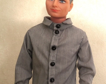 May include: A male doll with dark hair and blue eyes wearing a long-sleeved, grey and white pinstriped button-down shirt with a small collar and five black buttons down the front, paired with brown trousers that are visible at the waist.
