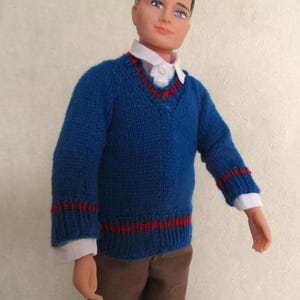May include: A doll with dark hair and blue eyes wearing a blue V-neck jumper with red striped trim at the collar, cuffs, and hem. Underneath the jumper is a white collared shirt with two visible buttons. The doll wears brown trousers.