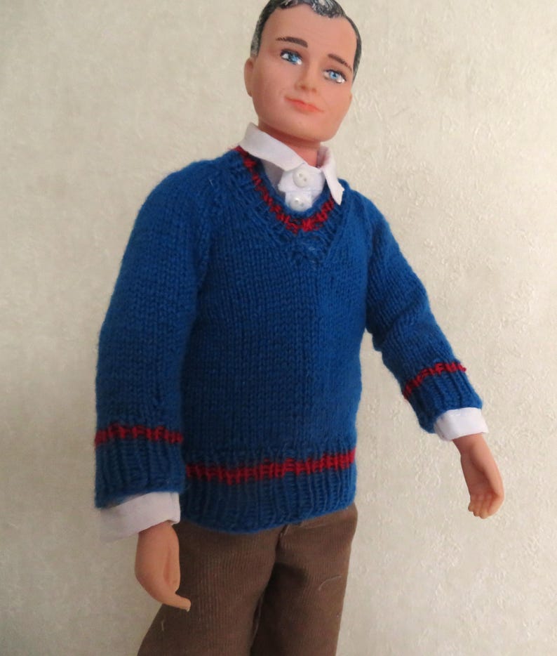 May include: A doll with dark hair and blue eyes wearing a blue V-neck jumper with red striped trim at the collar, cuffs, and hem. Underneath the jumper is a white collared shirt with two visible buttons. The doll wears brown trousers.