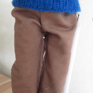 May include: A doll wearing a blue knitted jumper with a horizontal red stripe across the chest and solid brown trousers. The jumper features a ribbed texture and a crew neckline, while the trousers are made of a plain, smooth brown fabric.