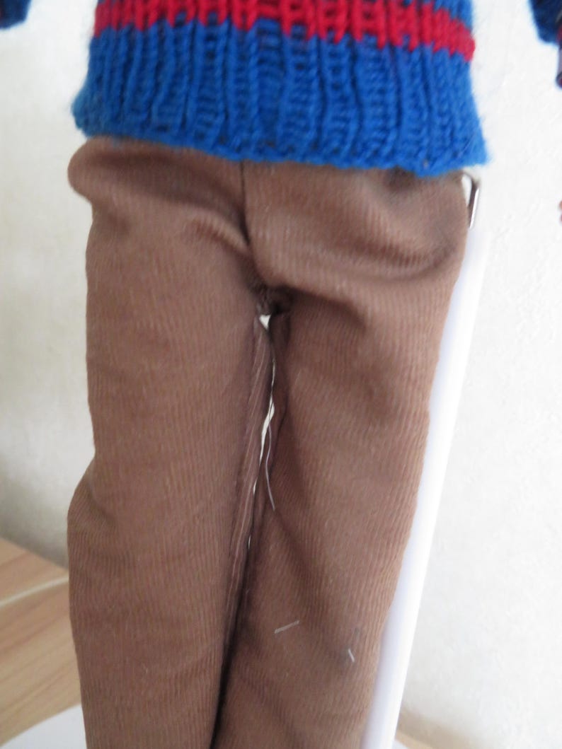 May include: A doll wearing a blue knitted jumper with a horizontal red stripe across the chest and solid brown trousers. The jumper features a ribbed texture and a crew neckline, while the trousers are made of a plain, smooth brown fabric.