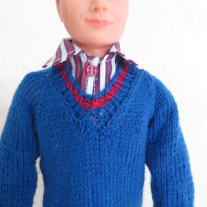 May include: A doll with moulded brown hair and blue eyes wearing a blue knitted V-neck jumper with red accents at the neckline and cuffs. Underneath the jumper is a button-down shirt featuring vertical stripes in white, navy blue, and red.