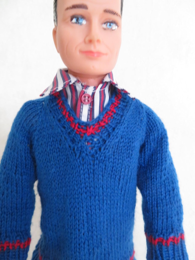 May include: A doll with moulded brown hair and blue eyes wearing a blue knitted V-neck jumper with red accents at the neckline and cuffs. Underneath the jumper is a button-down shirt featuring vertical stripes in white, navy blue, and red.