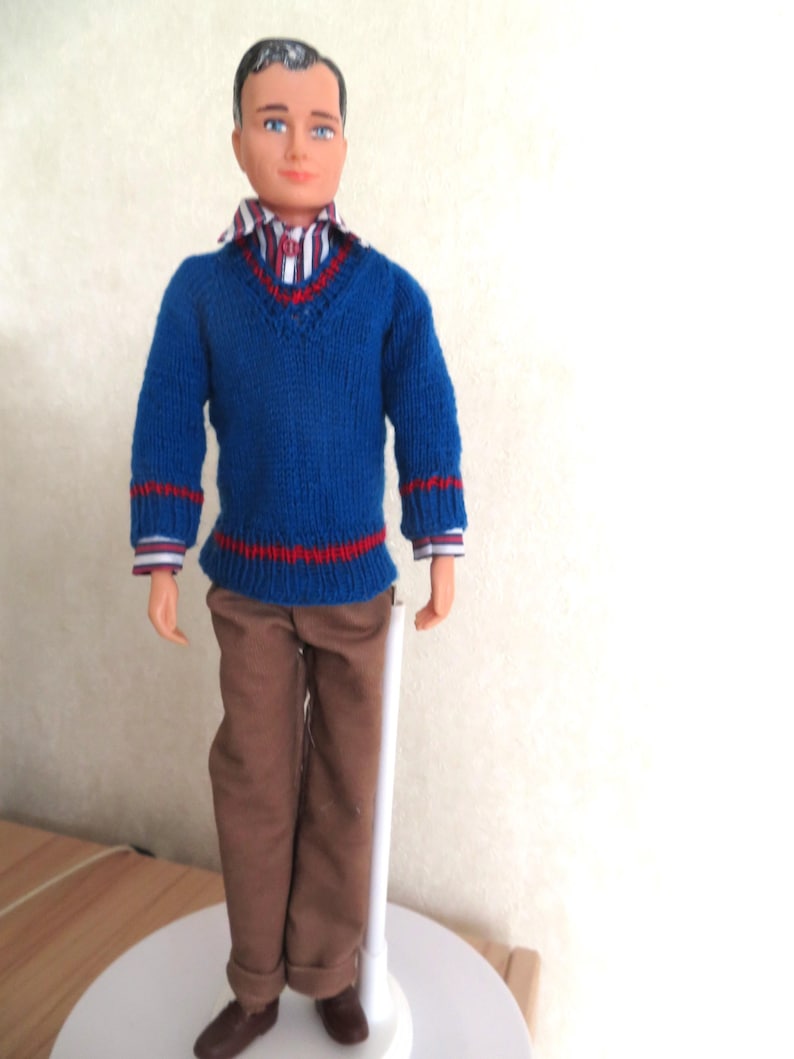 May include: A doll with dark hair and blue eyes wearing a blue V-neck jumper with red stripes on the cuffs and hem, a striped collared shirt underneath, and brown trousers. The doll is standing upright on a white circular base with a support stand.