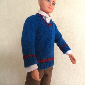 May include: A doll wearing a blue V-neck knit jumper with red stripes on the cuffs and hem, layered over a white collared shirt, paired with brown trousers. The doll has short dark hair, blue eyes, and a neutral expression on its face.