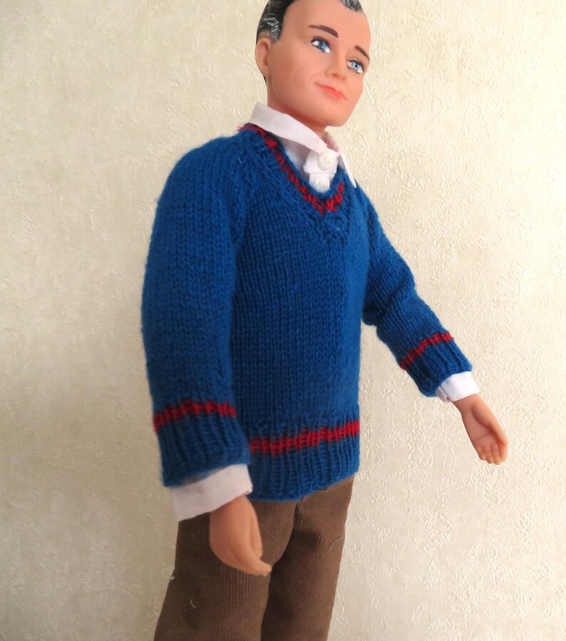May include: A doll wearing a blue V-neck knit jumper with red stripes on the cuffs and hem, layered over a white collared shirt, paired with brown trousers. The doll has short dark hair, blue eyes, and a neutral expression on its face.
