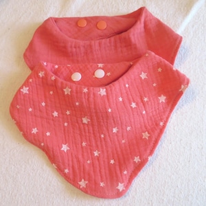 May include: Two coral-colored baby bibs. One bib is plain, and the other is patterned with white stars. Both bibs have snap closures. The bibs are made of a soft, textured fabric, and are likely made of muslin.
