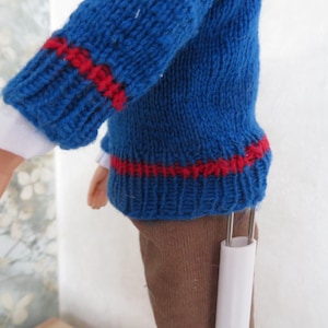 May include: A doll wearing a blue knitted jumper with red horizontal stripes on the sleeves and hem, layered over a white long-sleeved shirt. The doll is also wearing brown trousers and is supported by a white plastic stand.