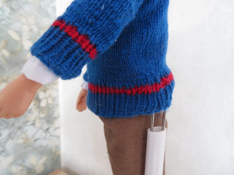 May include: A doll wearing a blue knitted jumper with red horizontal stripes on the sleeves and hem, layered over a white long-sleeved shirt. The doll is also wearing brown trousers and is supported by a white plastic stand.