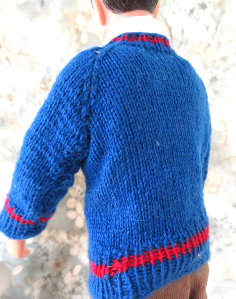 May include: A hand-knitted blue jumper with long sleeves, featuring a ribbed crew neckline and a ribbed hem. The jumper is accented with a thin horizontal red stripe around the collar, the cuffs of the sleeves, and the bottom hemline.