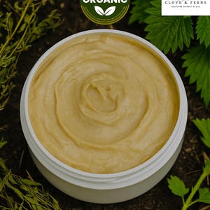 May include: A white jar filled with a light yellow cream. The cream has a smooth, swirled texture. The jar is surrounded by green leaves and brown soil. A green and white circular label reads "100% ORGANIC". The brand name "CLOVE & FERNS" is also visible.