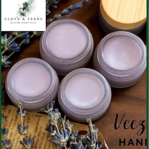 May include: Four small, clear jars filled with a light purple balm are arranged on a wooden surface, with dried lavender sprigs and a piece of aged paper. The jars have a silver rim and a light purple lid. The logo "Clove & Ferns" is in the upper left corner.
