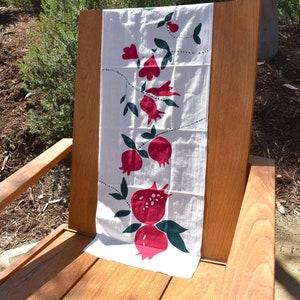 May include: A white cotton tea towel with a red and green pomegranate design. The towel is folded and placed on a wooden chair.