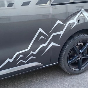 Könnte beinhalten: Ein dunkelgraues Fahrzeug mit einer weißen Bergketten-Grafik an der Seite. Das Fahrzeug hat einen schwarzen Reifen und eine Felge. Das Bergketten-Design besteht aus einer Reihe weißer, eckiger Linien.