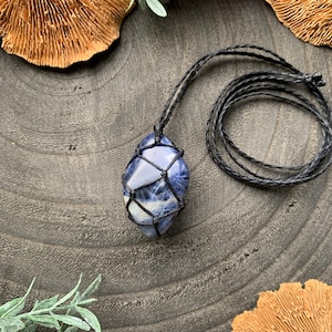 Puede incluir: Un collar con colgante de sodalita azul con un cordón negro tejido. La piedra está encerrada en una red negra. El collar se exhibe sobre una superficie gris oscura texturizada, rodeado de elementos decorativos.