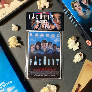 Może przedstawiać: Prostokątny magnes i etui na DVD z filmem "The Faculty". Magnes przedstawia tytuł filmu i twarze aktorów. Etui na DVD jest niebieskie z tytułem filmu. Popcorn jest rozrzucony.