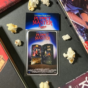 May include: A "Puppet Master" magnet with the movie poster design, featuring a cabinet with puppets. The magnet is in a clear plastic sleeve, surrounded by popcorn. The text "Evil Comes In All Sizes" and "Puppet Master" are visible.