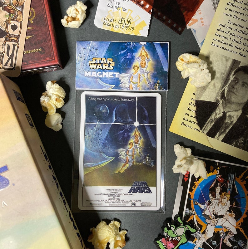 K&ouml;nnte beinhalten: Ein Star Wars-Magnet und ein Filmplakat werden mit Popcorn pr&auml;sentiert. Der Magnet zeigt das Star Wars-Logo und eine Filmszene. Das Poster tr&auml;gt den Text "Vor langer Zeit in einer weit, weit entfernten Galaxis." und das Star Wars-Logo.
