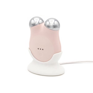 May include: A pink facial device with two silver metal rollers, charging on a white base with a white cord. The device has the word "nuFACE" printed on it.