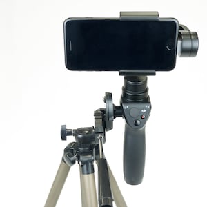 May include: A black smartphone gimbal mounted on a tripod. The gimbal has a black handle with a control button and a red light. The tripod is made of metal and has three legs.