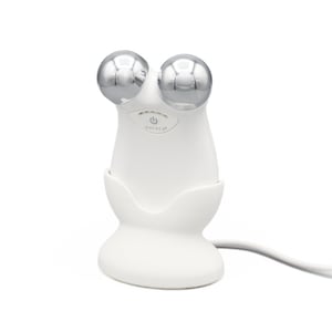 May include: White facial roller with two silver metal rollers, on a white charging base with a power cord. The device has the word "NuFACE" printed on it.