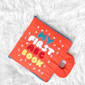 May include: Orange fabric book with a blue border and a handle. The book has the text "My First ABC Book" in colorful letters.