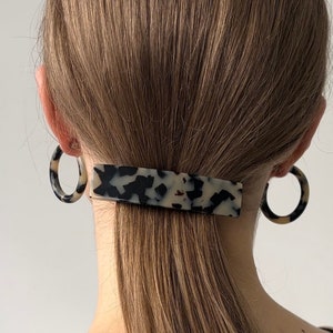 Peut inclure: Une pince à cheveux en écaille de tortue de forme rectangulaire, tenant une queue de cheval brune. La personne porte de grandes boucles d'oreilles rondes en écaille de tortue.