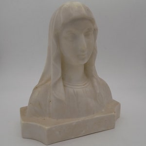 Op de afbeelding: Witte gipsen buste van een vrouw met haar hoofd naar beneden gekanteld. De buste staat op een rechthoekige basis met het woord "MATER" erop gegraveerd.