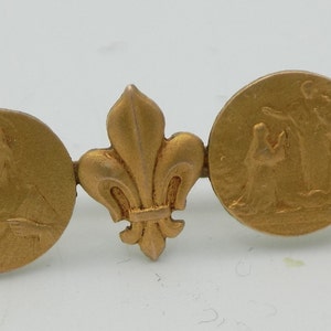 Puede incluir: Un pin de metal dorado con un diseño que presenta una flor de lis, un medallón redondo con una imagen en relieve de una persona y una flor de lis más pequeña.