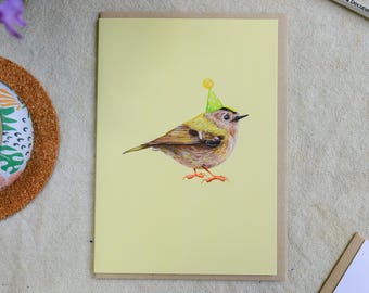 May include: A greeting card featuring a detailed illustration of a small bird wearing a party hat. The bird is depicted with brown, yellow, and green feathers, standing on a light yellow background. The card is on a neutral surface with paintbrushes and flowers.
