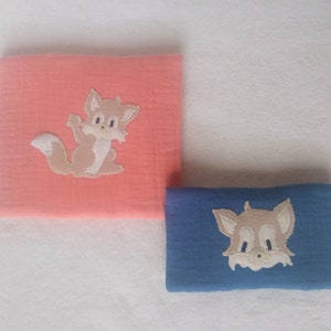 Könnte beinhalten: Zwei weiche, aus Baumwolle gefertigte Musselin-Tücher mit gestickten Fuchs-Motiven. Ein Tuch ist pfirsichfarben mit einem braunen Fuchs, das andere ist blau mit einem braunen Fuchs.