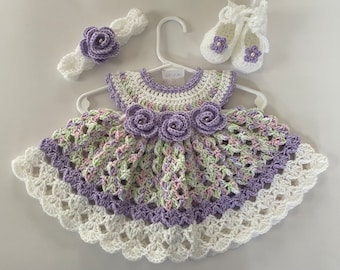 May include: A crocheted baby dress with a lavender and green color scheme. The dress has a white crocheted trim and a matching headband with a lavender flower. The dress also comes with a pair of white crocheted shoes with lavender flowers.