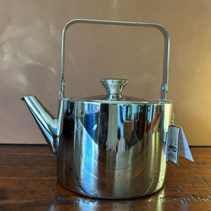May include: A stainless steel teapot with a handle and lid. The teapot has a spout and a tag attached to it. The teapot is sitting on a wooden surface.