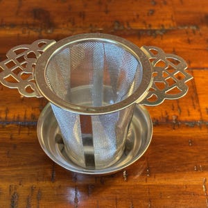 May include: A stainless steel tea infuser with a fine mesh basket and decorative handles. The infuser sits in a small, matching saucer. The handles have an ornate, openwork design. The tea infuser is designed for steeping loose leaf tea.