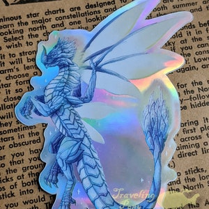 May include: A holographic sticker featuring a blue dragon with a white background. The dragon is in a standing position with its wings spread out. The sticker has a holographic finish that shimmers in different colours. The sticker is a great addition to any dragon lover's collection.