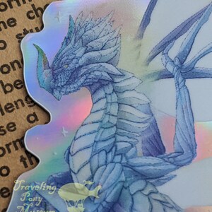 May include: A holographic sticker featuring a blue dragon with iridescent wings. The dragon is facing the left side of the image. The sticker has a white background with a faint rainbow sheen. The sticker has the text "Traveling Pony Museum" and "www.etsy.com/shop/LetYourArtTravel" at the bottom.