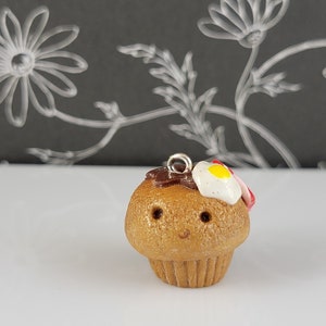 May include: A small, brown, clay muffin with a white and yellow egg and red bacon topping. The muffin has a smiley face and a silver loop for hanging.