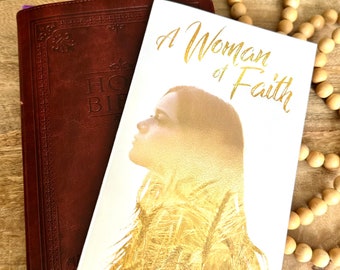 May include: A white book with the title "A Woman of Faith" in gold lettering. The cover features a silhouette of a woman with a golden background. The book is titled "Girlfriends in the Word" by Natalie Dinnim.