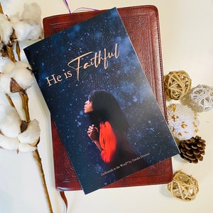 May include: A brown leather-bound book with a bookmark. The book is open to a page with a woman praying and the words "He is Faithful" in gold lettering. The book is titled "Girlfriends in the Word" by Natalie Drumm.