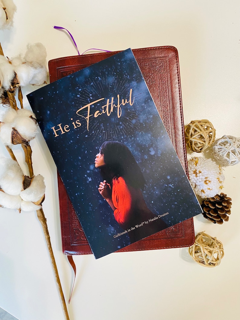 May include: A brown leather-bound book with a bookmark. The book is open to a page with a woman praying and the words "He is Faithful" in gold lettering. The book is titled "Girlfriends in the Word" by Natalie Drumm.