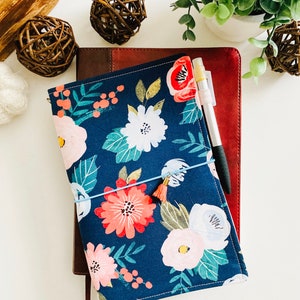 May include: A blue floral notebook cover with a pink and white floral pattern. The cover is wrapped around a brown leather notebook. A pen is placed on top of the notebook.