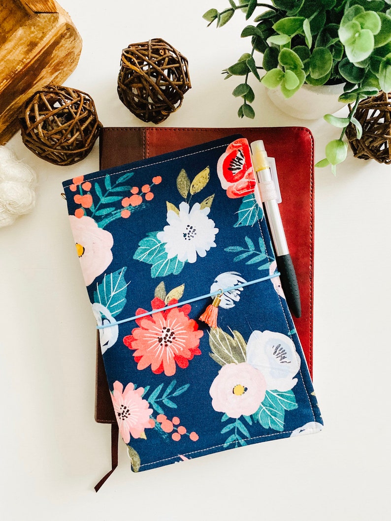 May include: A blue floral notebook cover with a pink and white floral pattern. The cover is wrapped around a brown leather notebook. A pen is placed on top of the notebook.